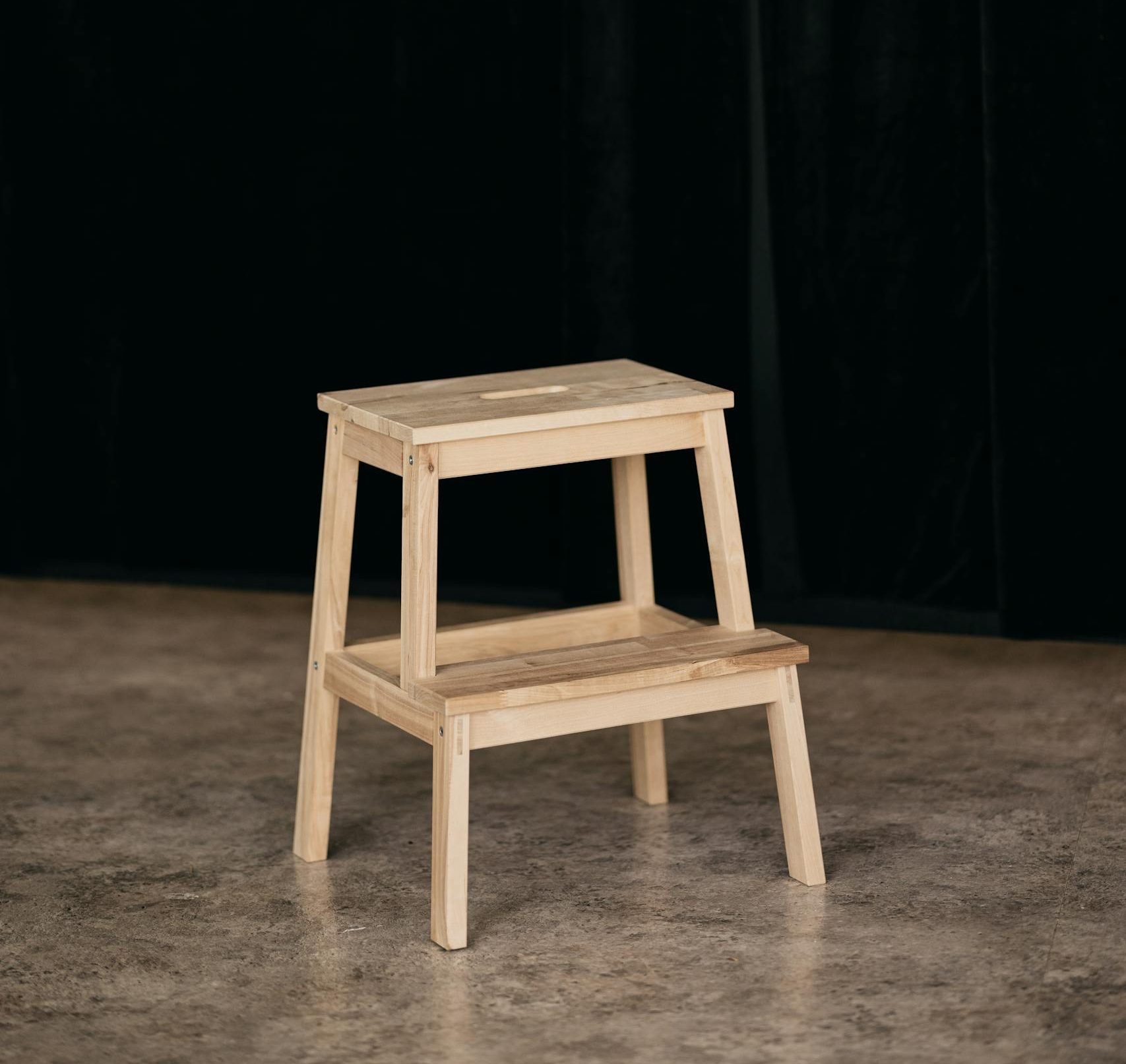 A simple wooden step stool in front of a black curtain on a stone floor.