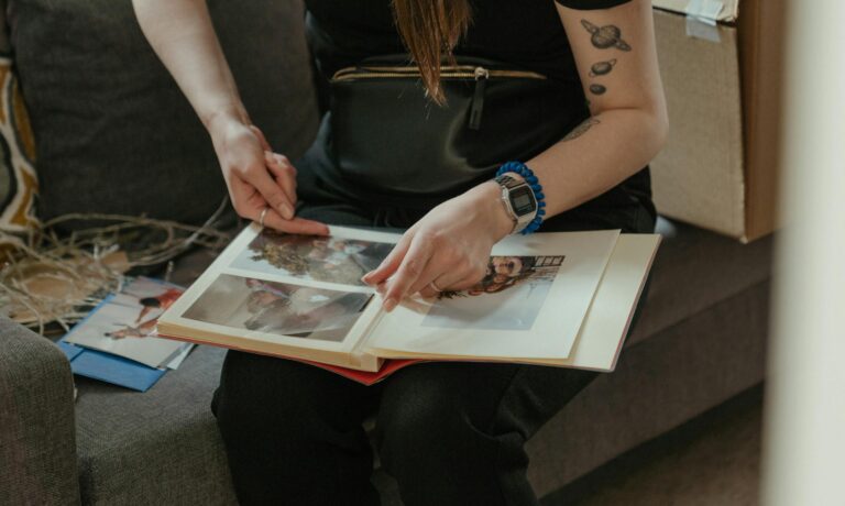 A person with tattoos sits indoors examining a photo album on a sofa.
